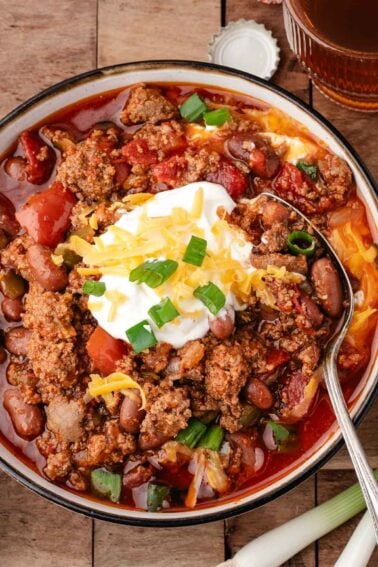 Bowl of chili with ground meat, beans, and tomatoes, topped with shredded cheese, sour cream, and chopped green onions, with a spoon resting in the bowl.