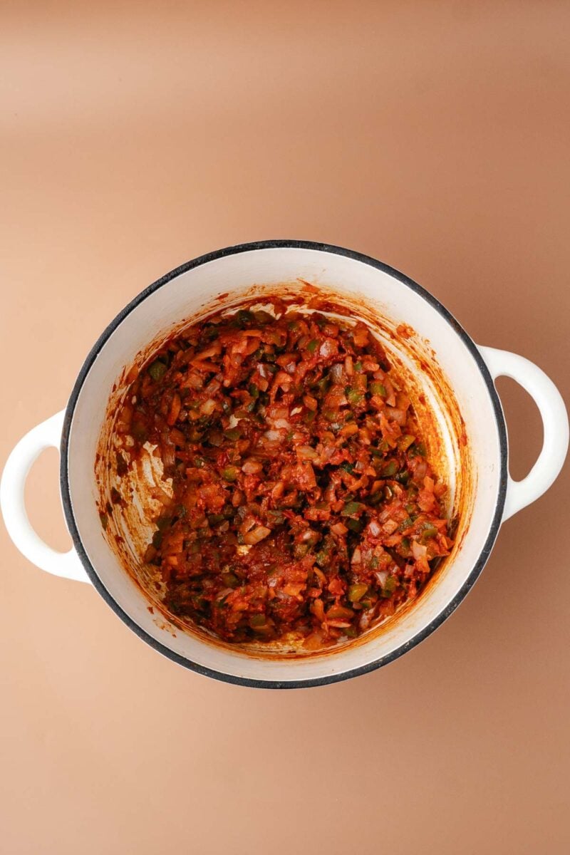 White pot containing a mixture of chopped onions, green bell peppers, and tomatoes, cooked down and seasoned, on a beige background.