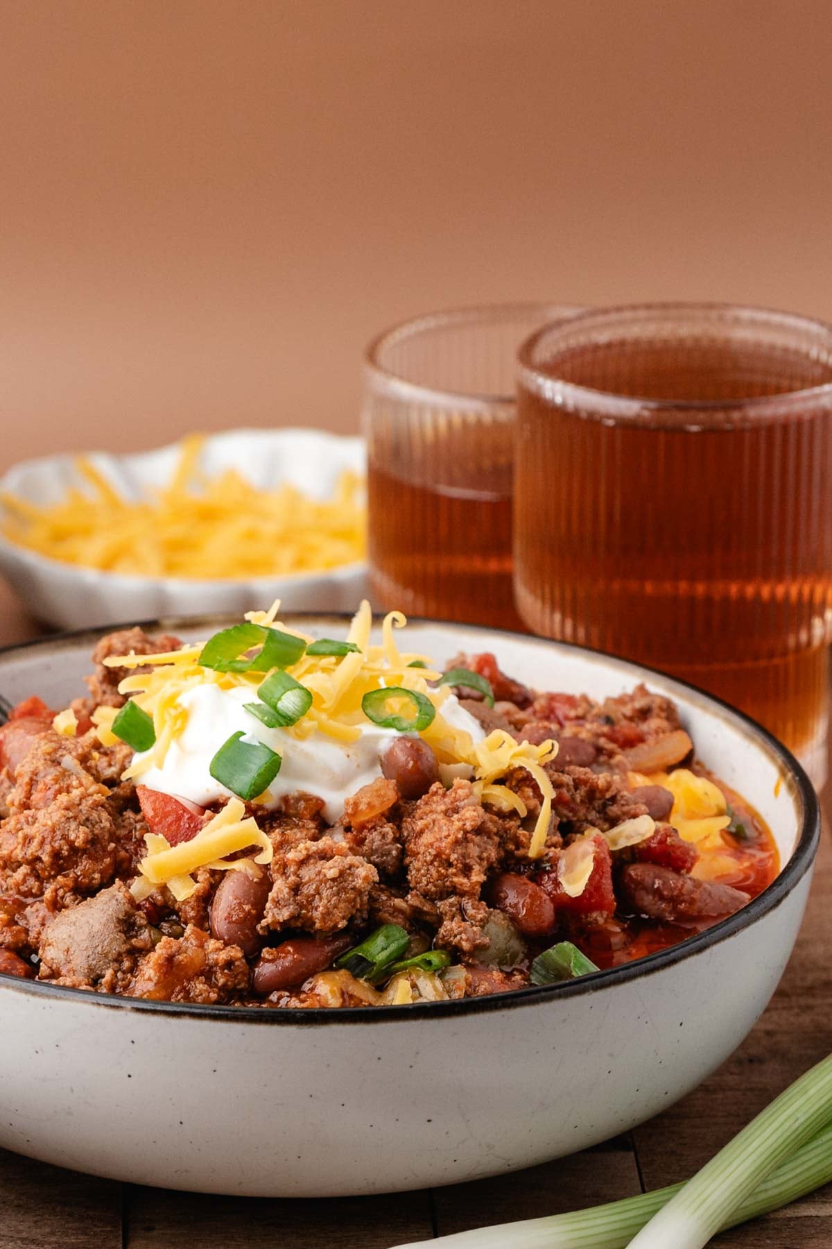 A bowl of chili topped with sour cream, shredded cheese, and green onions, with drinks and a bowl of shredded cheese in the background.