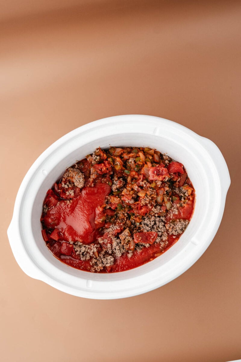White slow cooker filled with ground beef, chopped vegetables, and tomato sauce, on a light brown surface.