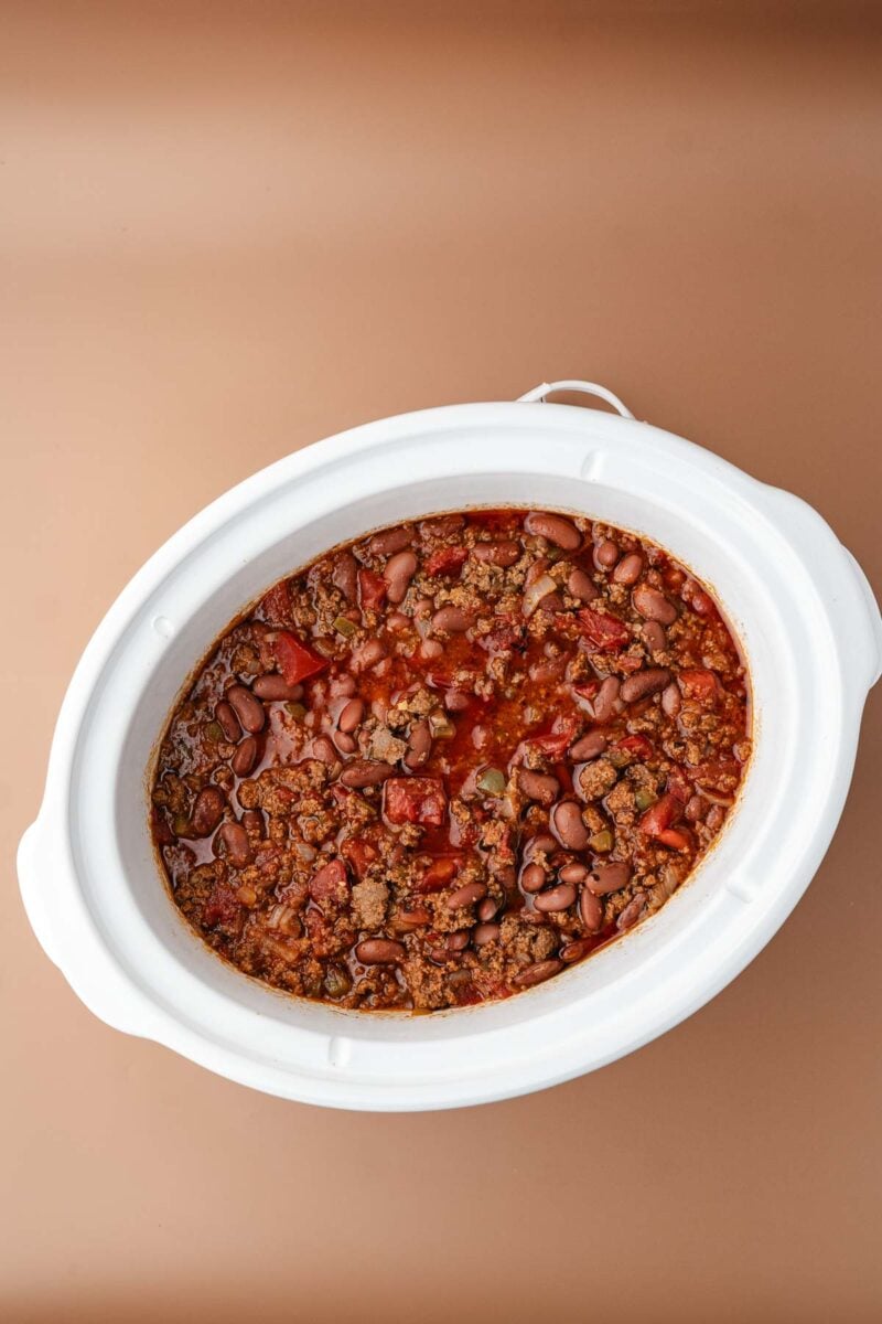 A white slow cooker filled with chili containing ground meat, red beans, and tomatoes, set against a light brown background.