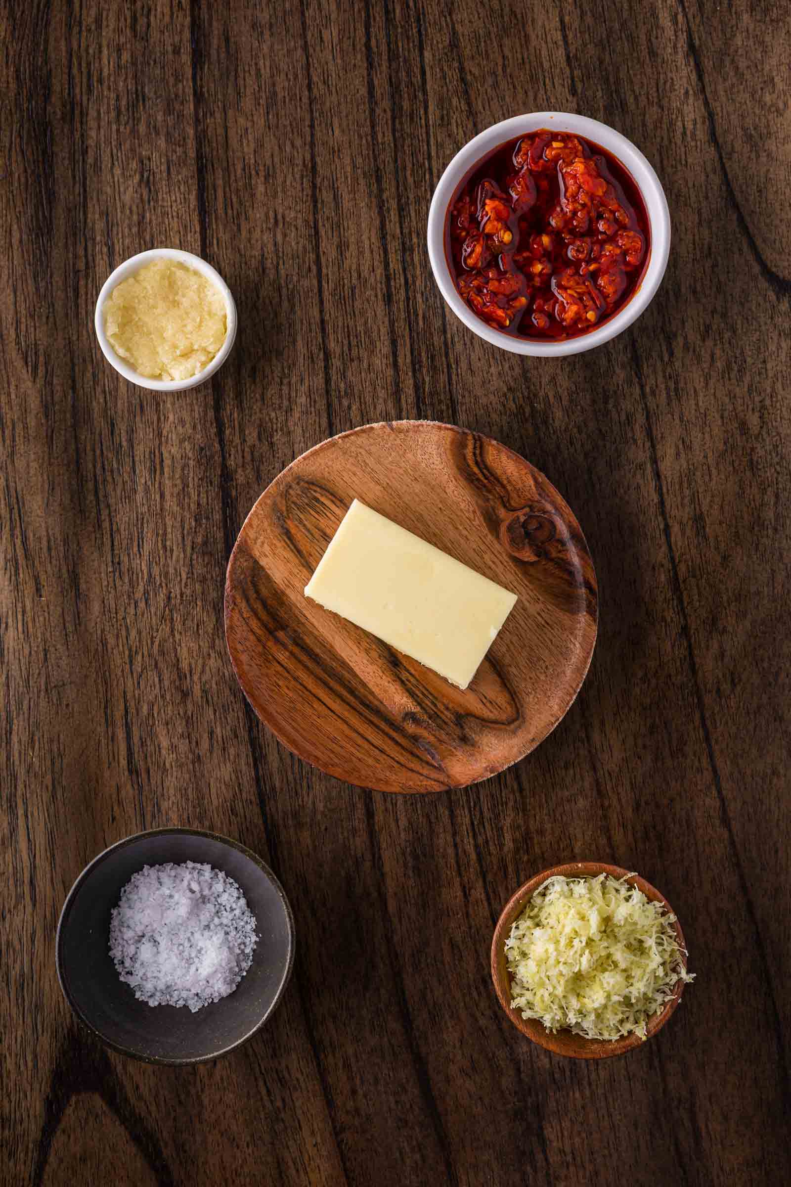 A wooden table with dishes containing a slab of butter, dried tomatoes, grated cheese, minced garlic, and coarse salt arranged in a circular pattern.