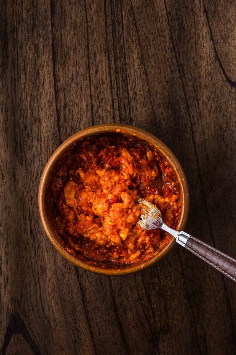 A wooden bowl filled with chili compound butter small metal spoon resting inside, sits on a dark wooden surface.