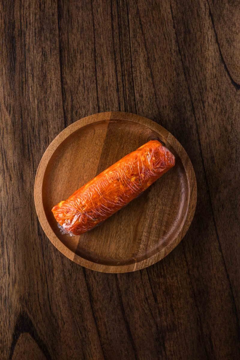 Chili compound butter, wrapped in plastic, sits on a round wooden plate atop a dark wooden surface.