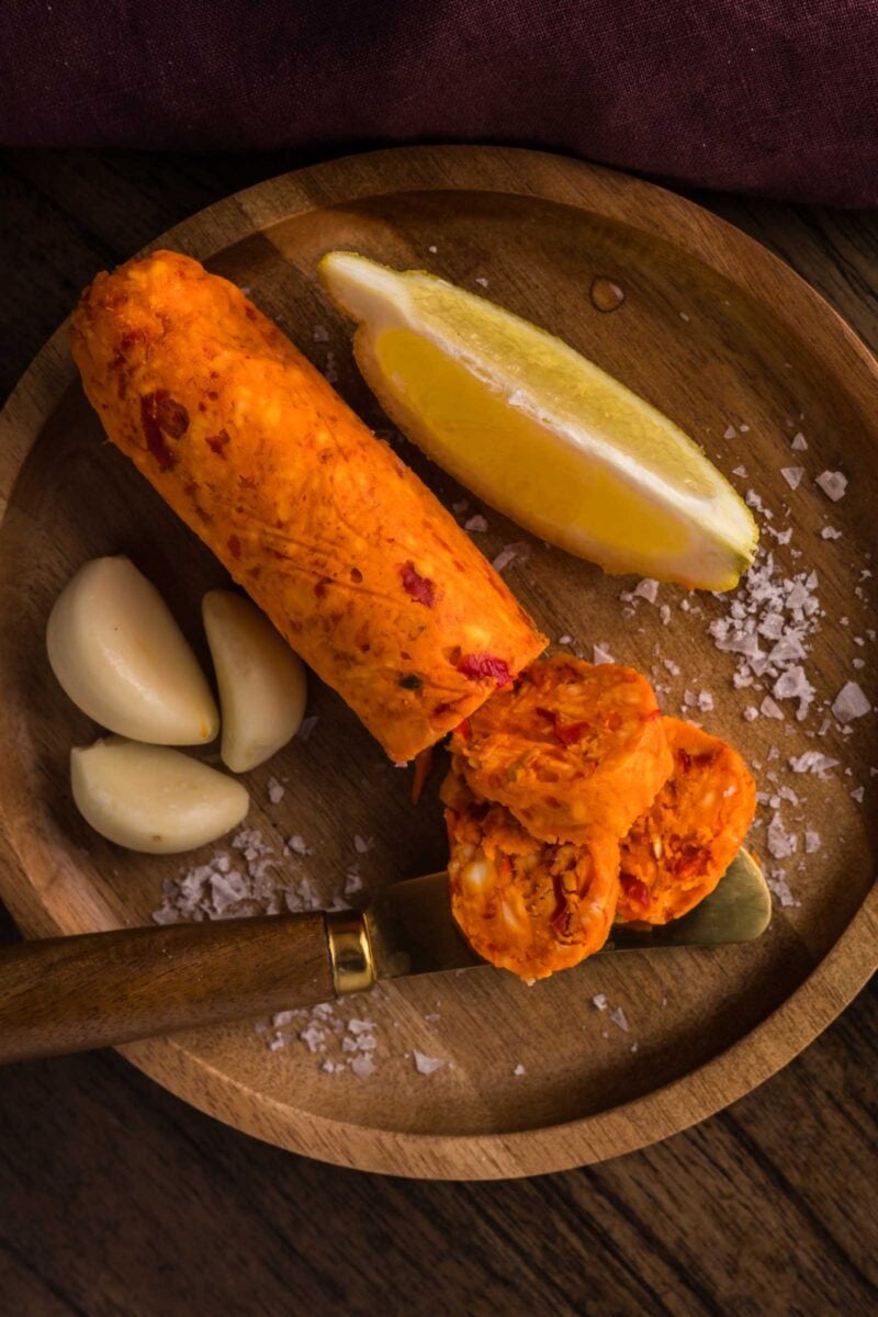 A wooden plate with a log of orange chili compound butter, three garlic cloves, a lemon wedge, coarse salt, and a knife.