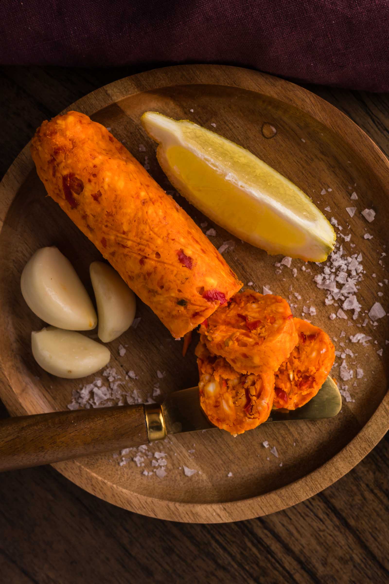 A wooden plate with a log of orange chili compound butter, three garlic cloves, a lemon wedge, coarse salt, and a knife.