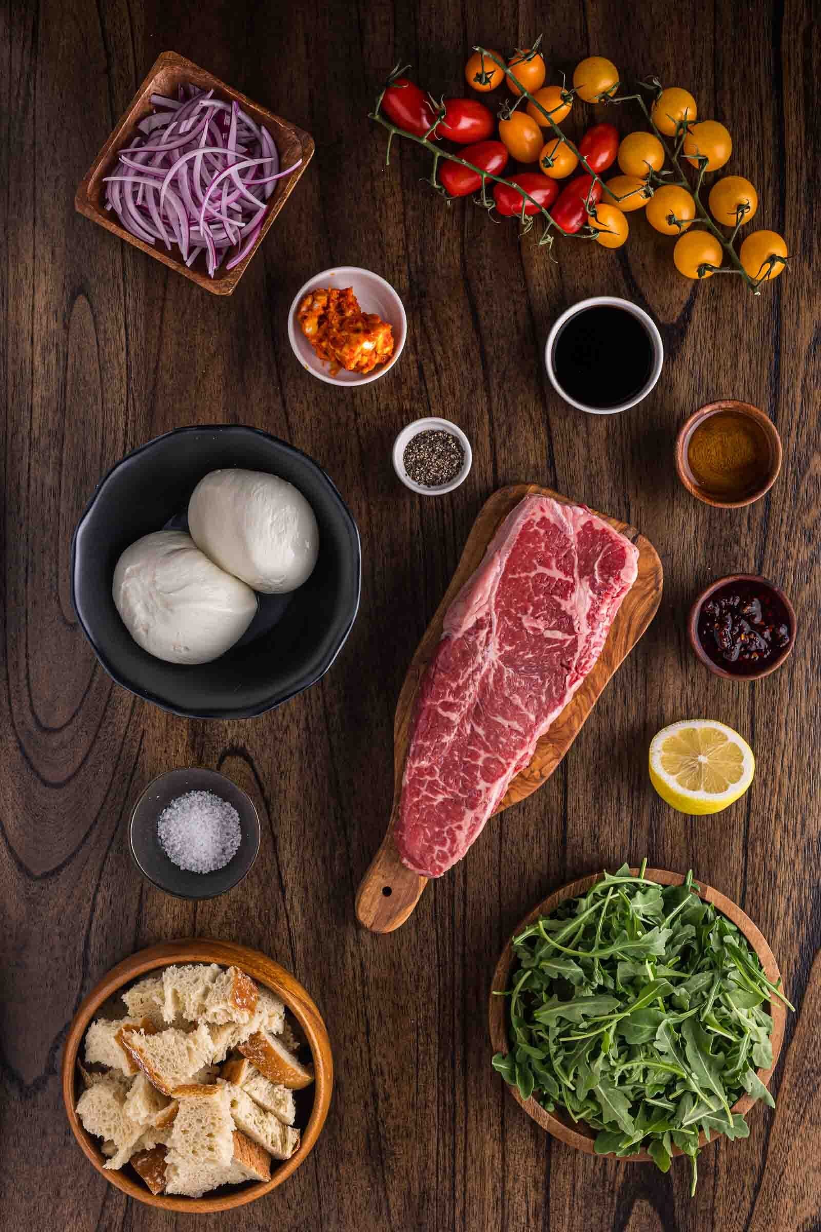 Steak, burrata, onions, cherry tomatoes, arugula, bread, lemon, salt, pepper, sauce, and condiments arranged on a wooden table.