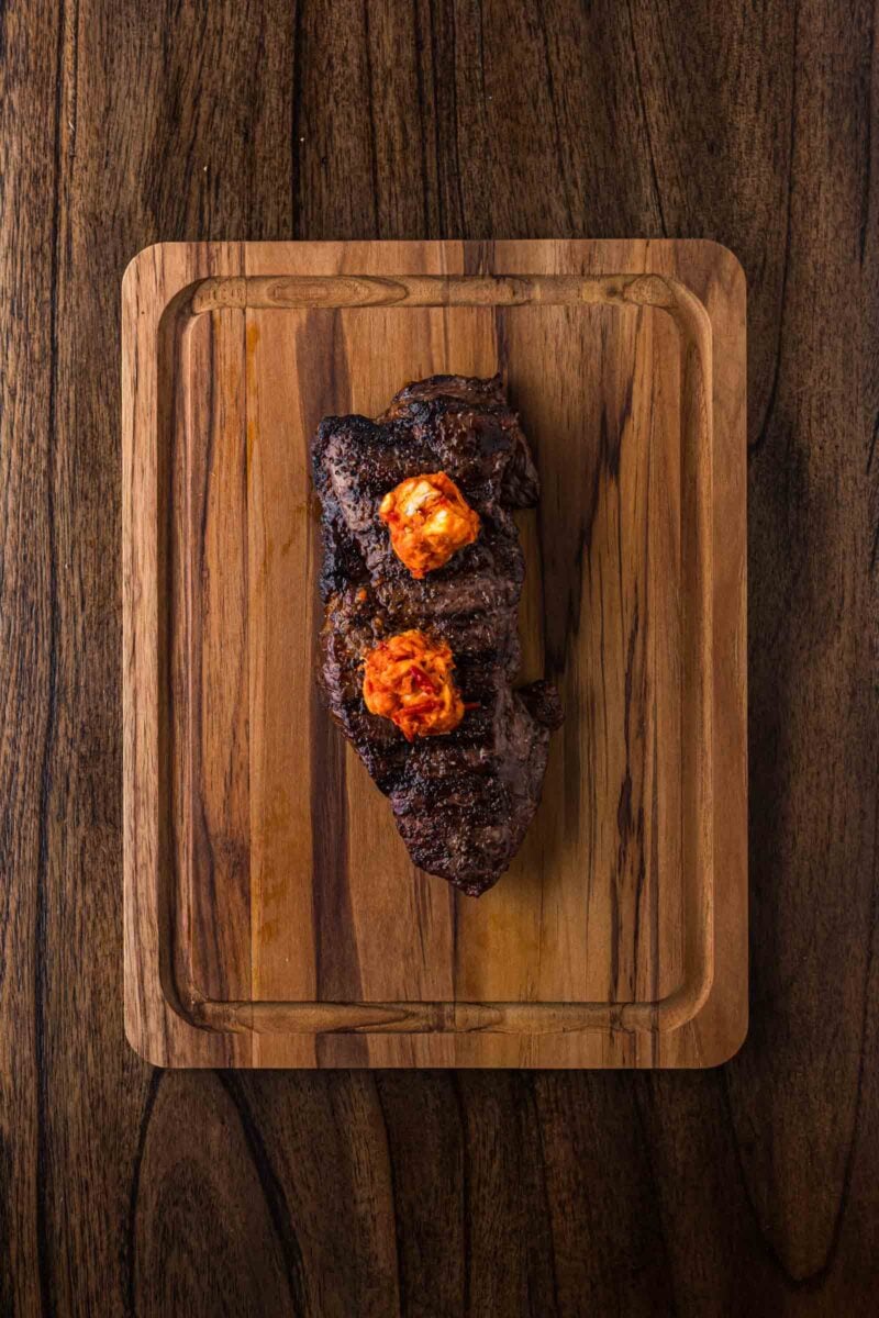 Grilled steak with two dollops of seasoned butter on top, served on a wooden cutting board, photographed from above.
