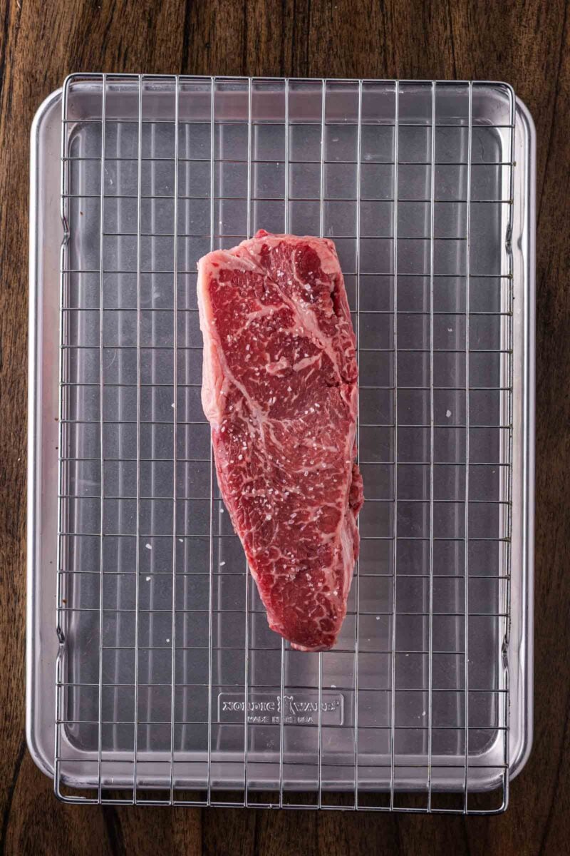 A raw steak sprinkled with salt is placed on a metal wire rack over a baking sheet, set on a wooden surface.