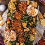Platter with sliced steak, leafy greens, burrata cheese, croutons, cherry tomatoes, red onion, and lemon halves, surrounded by small bowls of seasonings and vegetables on a wooden table.