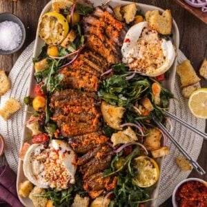 Platter with sliced steak, leafy greens, burrata cheese, croutons, cherry tomatoes, red onion, and lemon halves, surrounded by small bowls of seasonings and vegetables on a wooden table.
