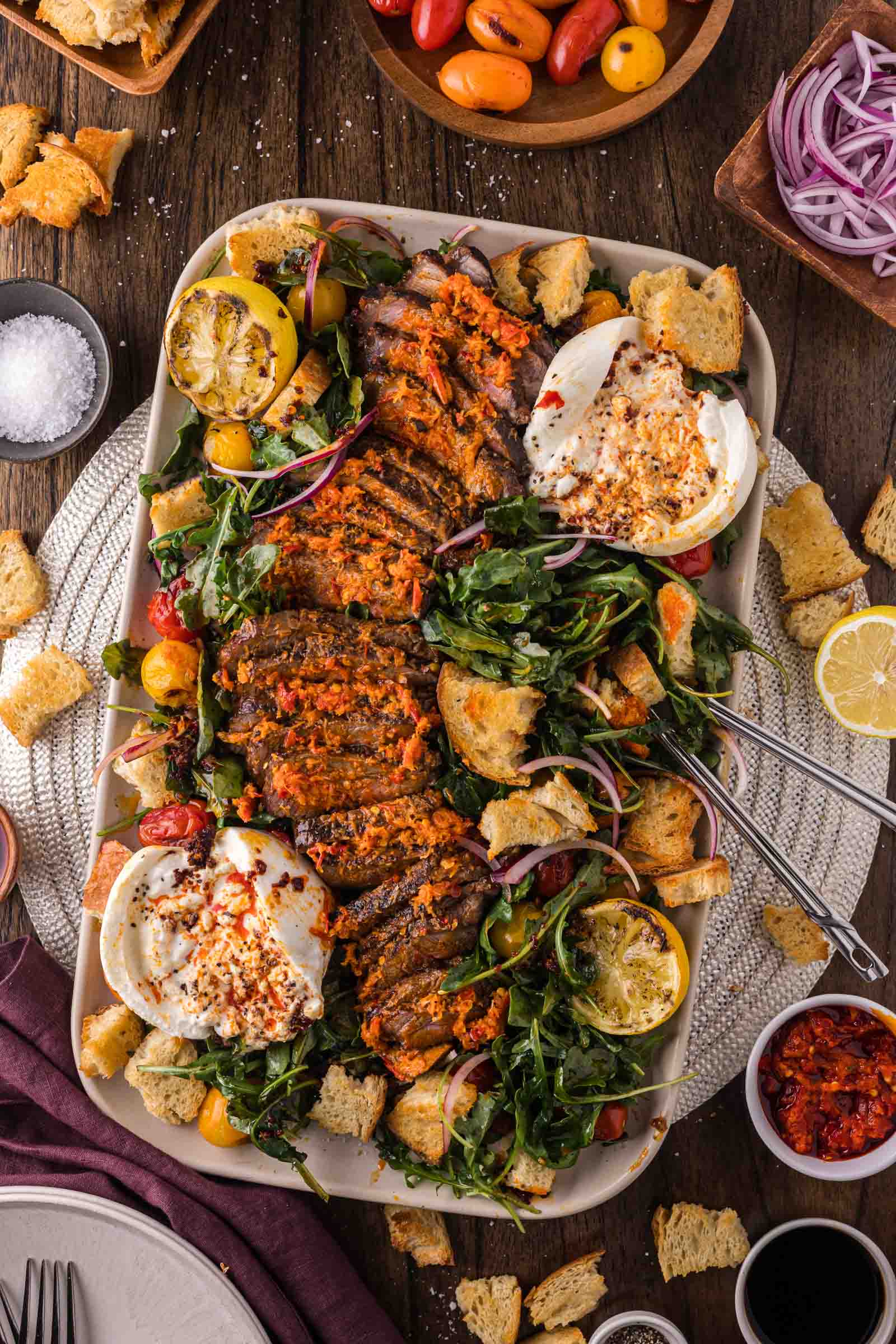 Platter with sliced steak, leafy greens, burrata cheese, croutons, cherry tomatoes, red onion, and lemon halves, surrounded by small bowls of seasonings and vegetables on a wooden table.