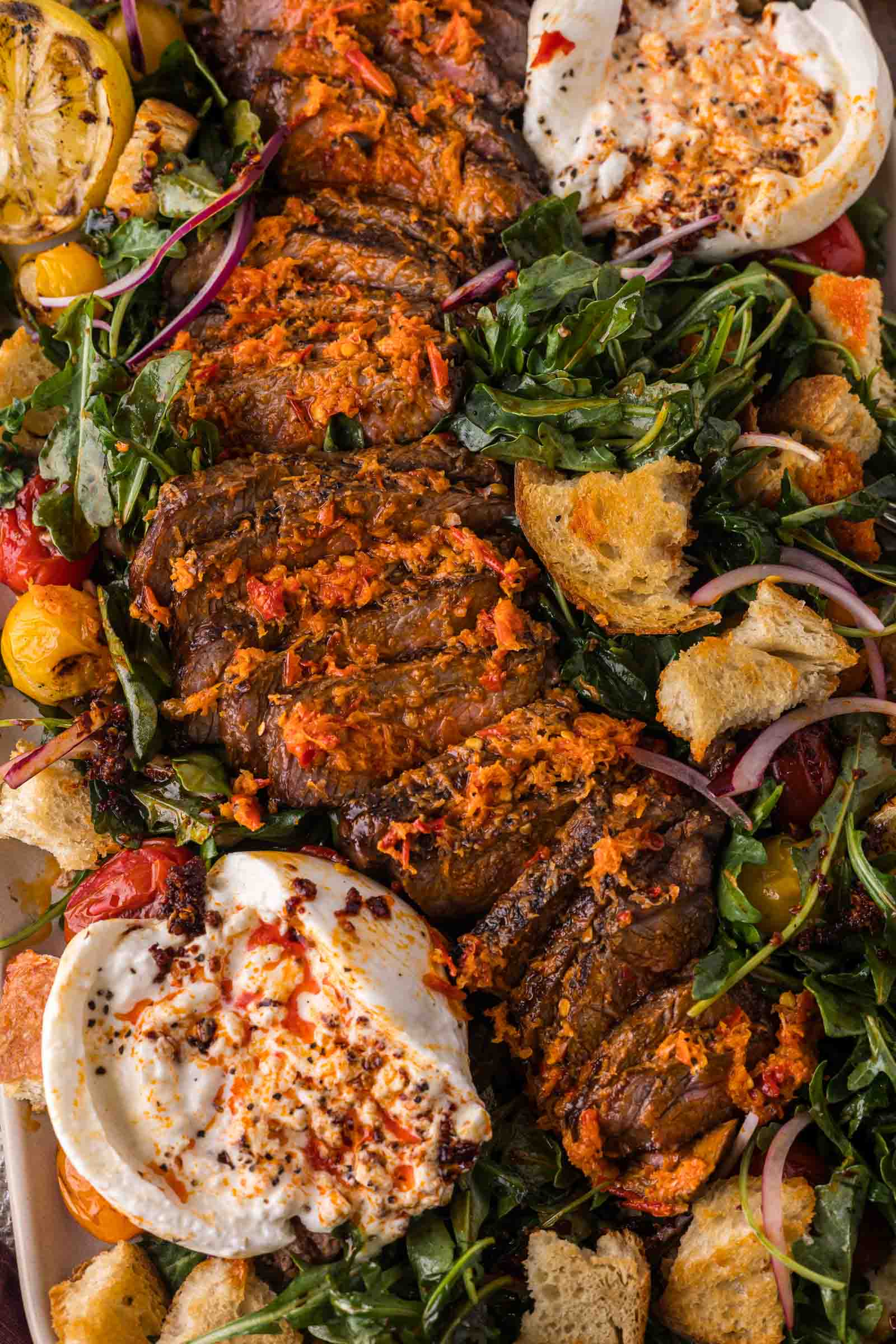 Sliced steak topped with melted chili butter on a bed of arugula, tomatoes, red onion, croutons, and two portions of burrata cheese.