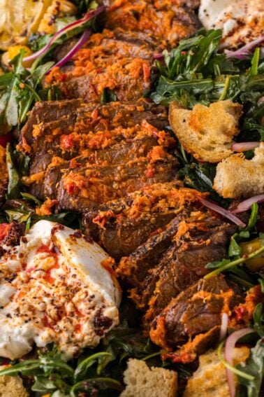 Sliced grilled steak on a bed of mixed greens, croutons, and dollops of burrata cheese with a red pepper topping.