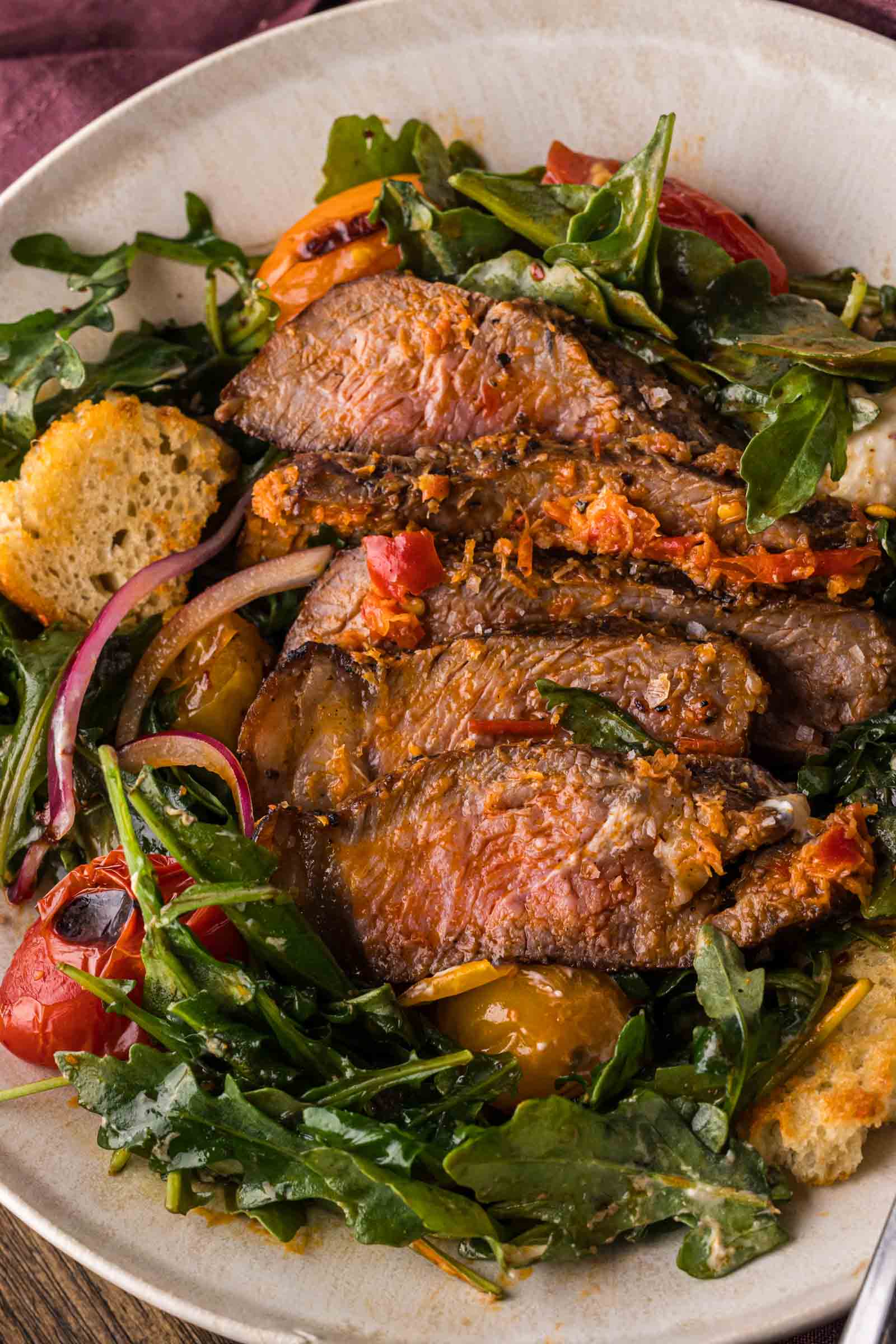 Sliced steak served on a bed of arugula with tomatoes, red onions, croutons, and dressing in a white bowl.