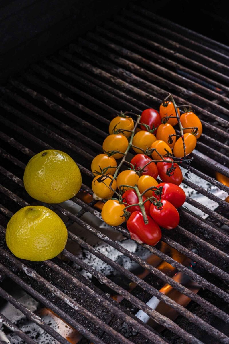 Two halved lemons and a cluster of yellow and red cherry tomatoes are placed on a grill with visible flames underneath.