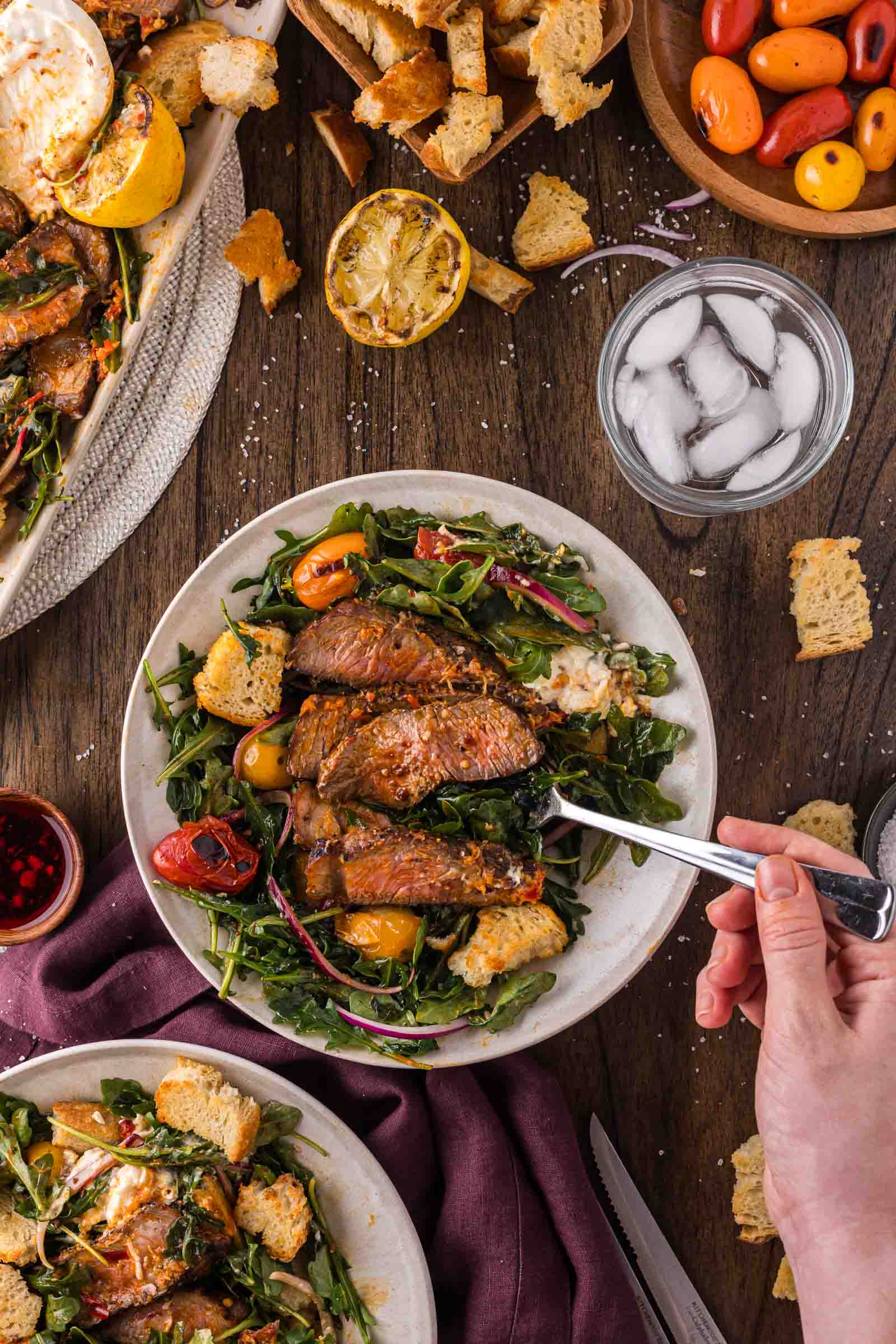 A plate of salad with grilled steak slices, cherry tomatoes, croutons, and greens on a wooden table, surrounded by bread, lemon halves, and a glass of ice water.