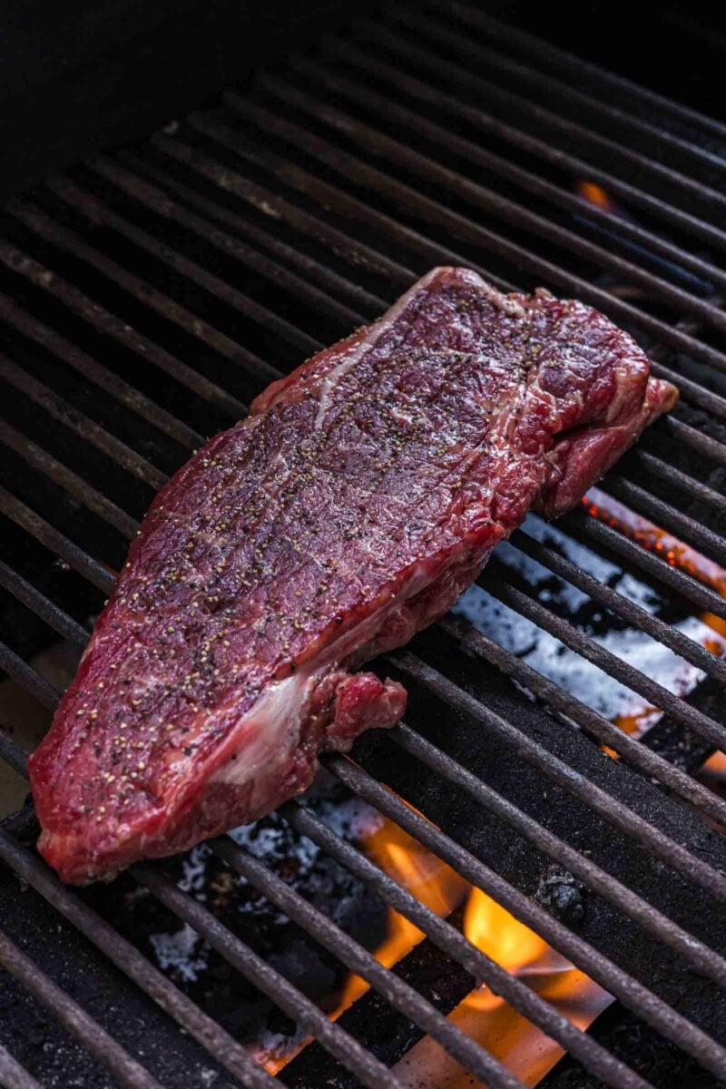 A seasoned raw steak is placed on a grill with flames visible below the grates.