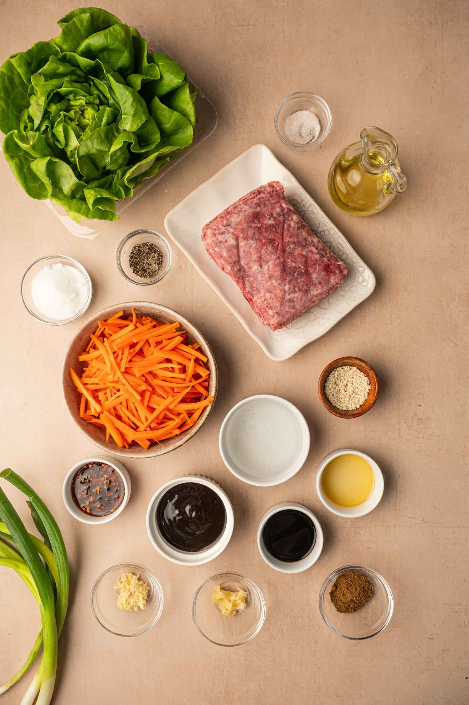 Ingredients for lamb five spice lettuce cups arranged on a beige surface, including ground meat, lettuce, shredded carrots, sauces, spices, green onion, oil, salt, and pepper in bowls and dishes.