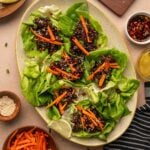A platter of five-spice lettuce wraps filled with a saucy sticky ground lamb and topped with julienned carrots, surrounded by bowls of carrot sticks, sesame seeds, lime wedges, and sauce.