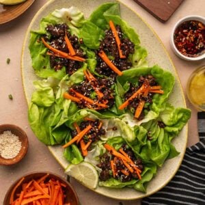 A platter of five-spice lettuce wraps filled with a saucy sticky ground lamb and topped with julienned carrots, surrounded by bowls of carrot sticks, sesame seeds, lime wedges, and sauce.