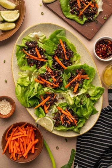 A platter of five-spice lettuce wraps filled with a saucy sticky ground lamb and topped with julienned carrots, surrounded by bowls of carrot sticks, sesame seeds, lime wedges, and sauce.