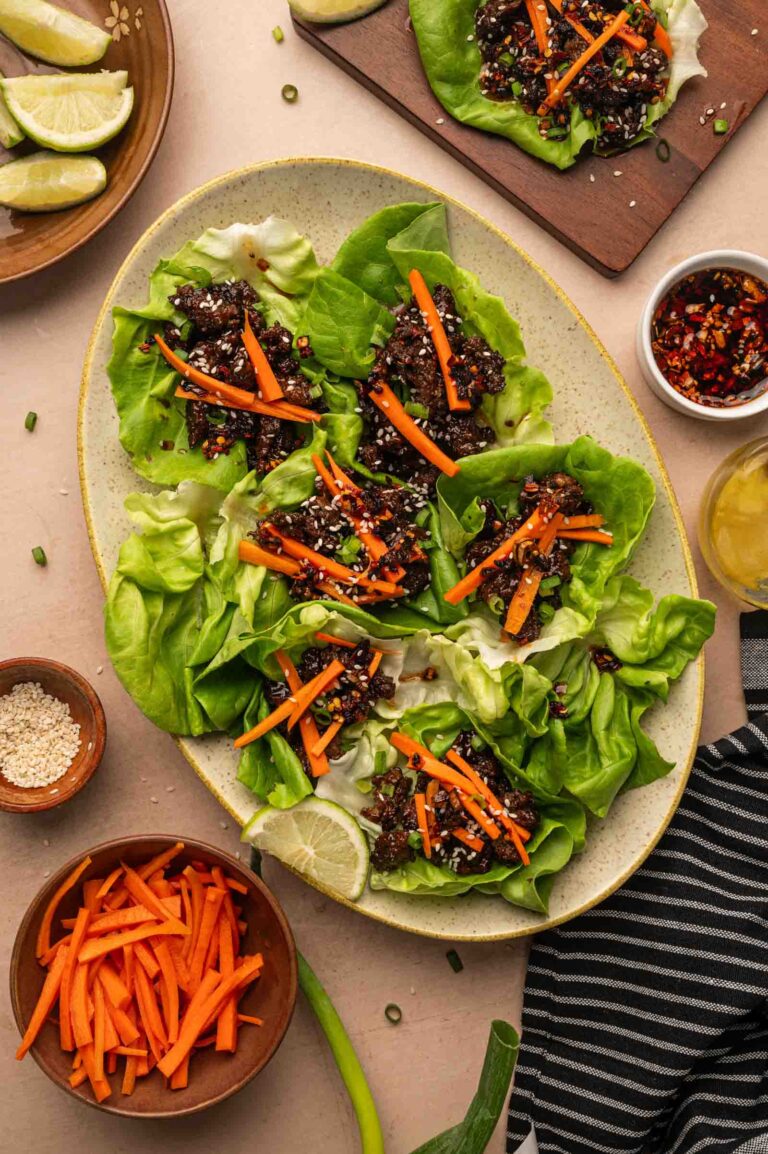 A platter of five-spice lettuce wraps filled with a saucy sticky ground lamb and topped with julienned carrots, surrounded by bowls of carrot sticks, sesame seeds, lime wedges, and sauce.
