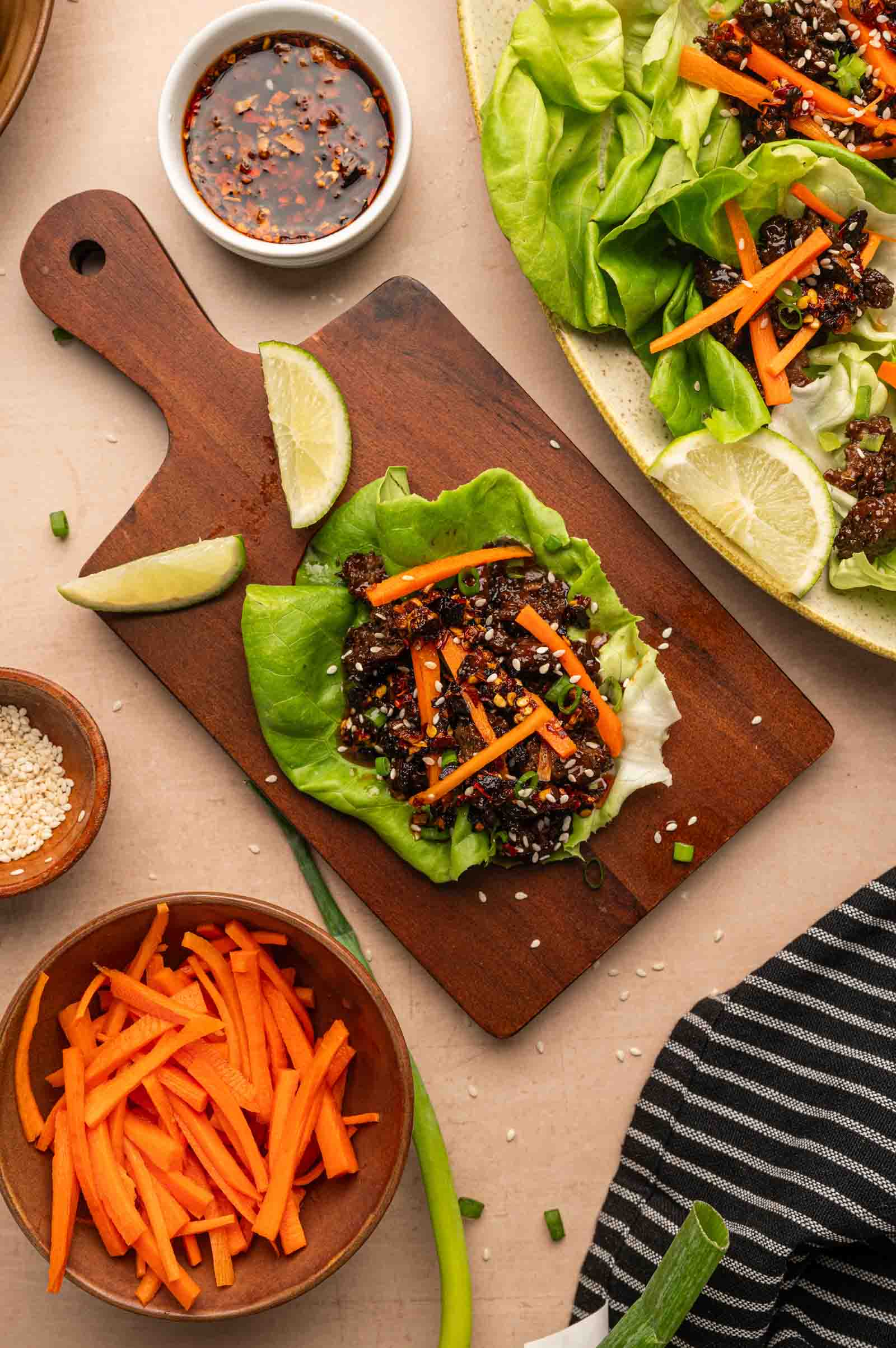 Lettuce cups with carrots and saucy ground lamb on a wooden board, surrounded by lime wedges, sauce, sesame seeds, and more carrot sticks.