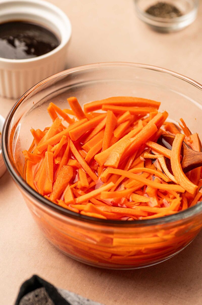 A glass bowl filled with julienned carrots is on a table. In the background, there is a small ramekin with a dark sauce and a small glass container with seasoning.