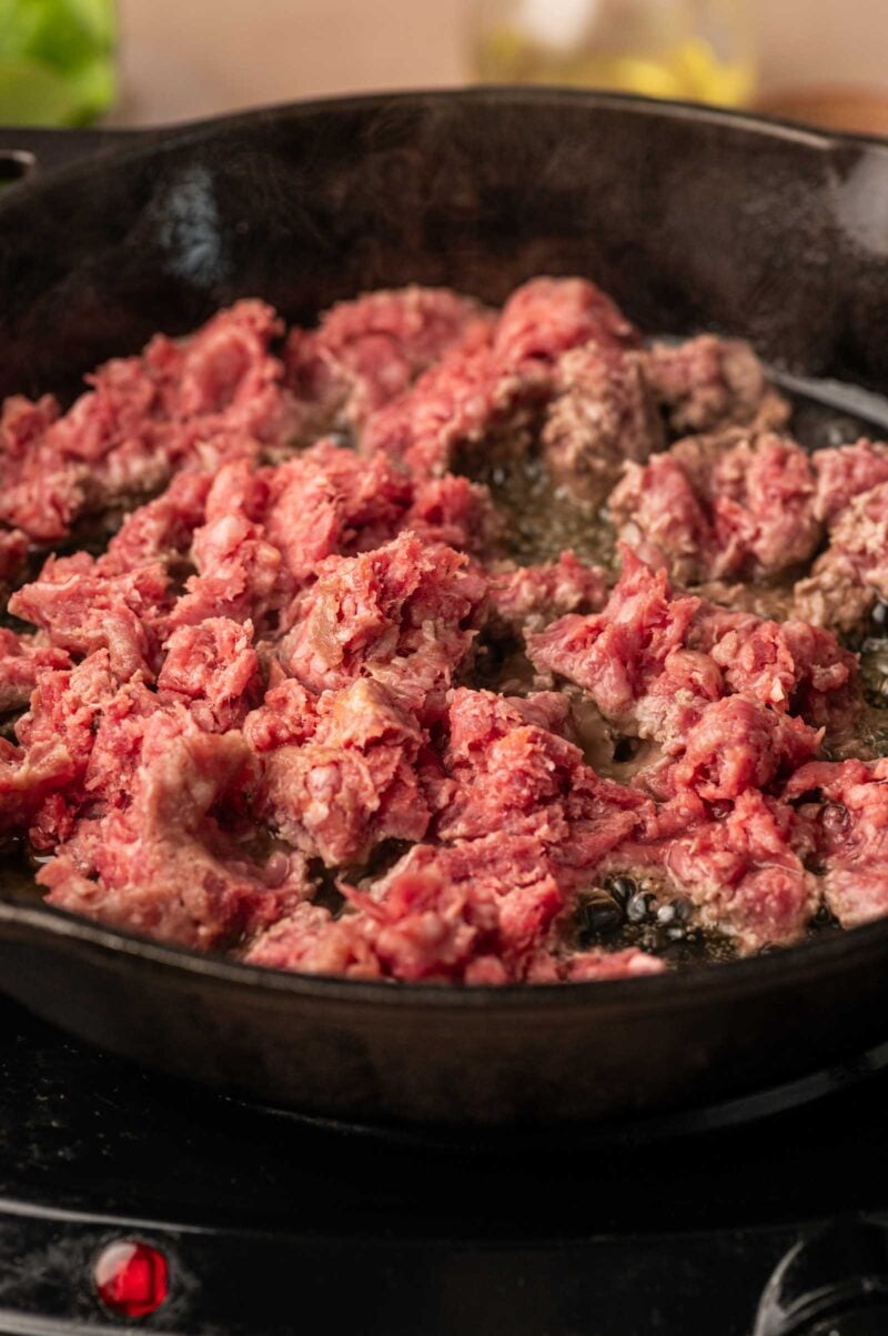 Ground lamb cooking in a cast iron skillet on a stovetop, with some pieces still pink and others starting to brown.