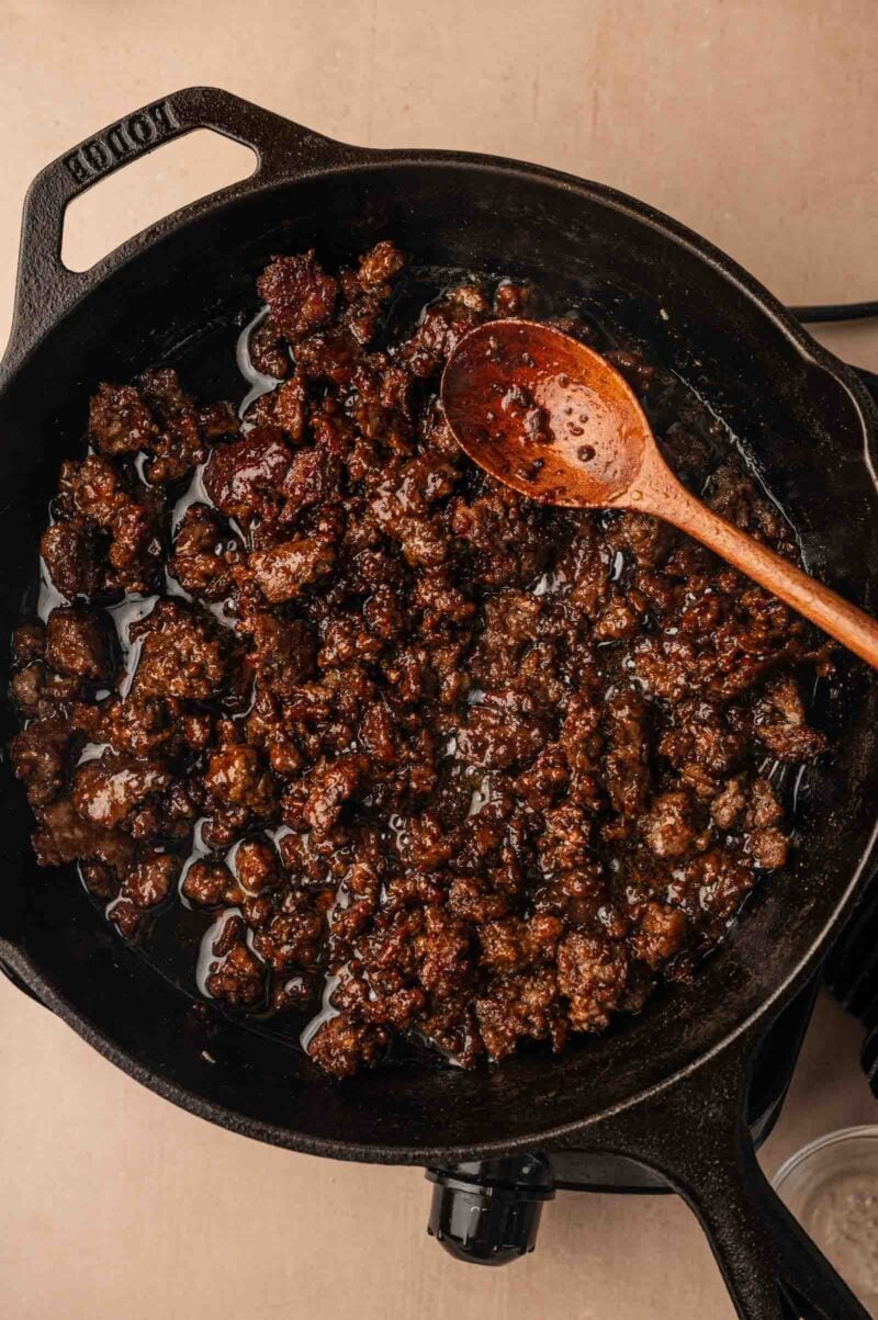 Ground lamb cooked in a cast iron skillet with a wooden spoon resting on the side, some oil visible at the bottom of the pan.