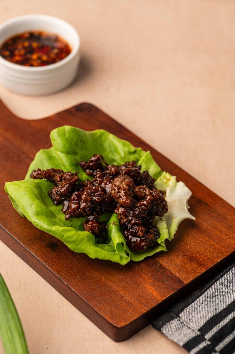 Cooked five spice ground lamb on a green lettuce leaf, served on a wooden board, with a small bowl of dark dipping sauce in the background.