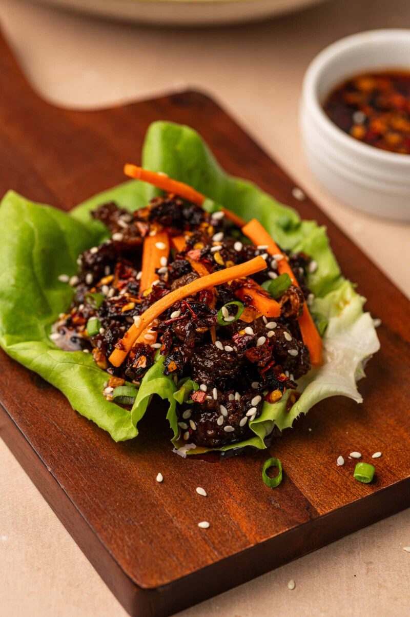 A lettuce cup topped with five spice ground lamb, shredded carrots, green onions, sesame seeds, and a small bowl of dipping sauce in the background.