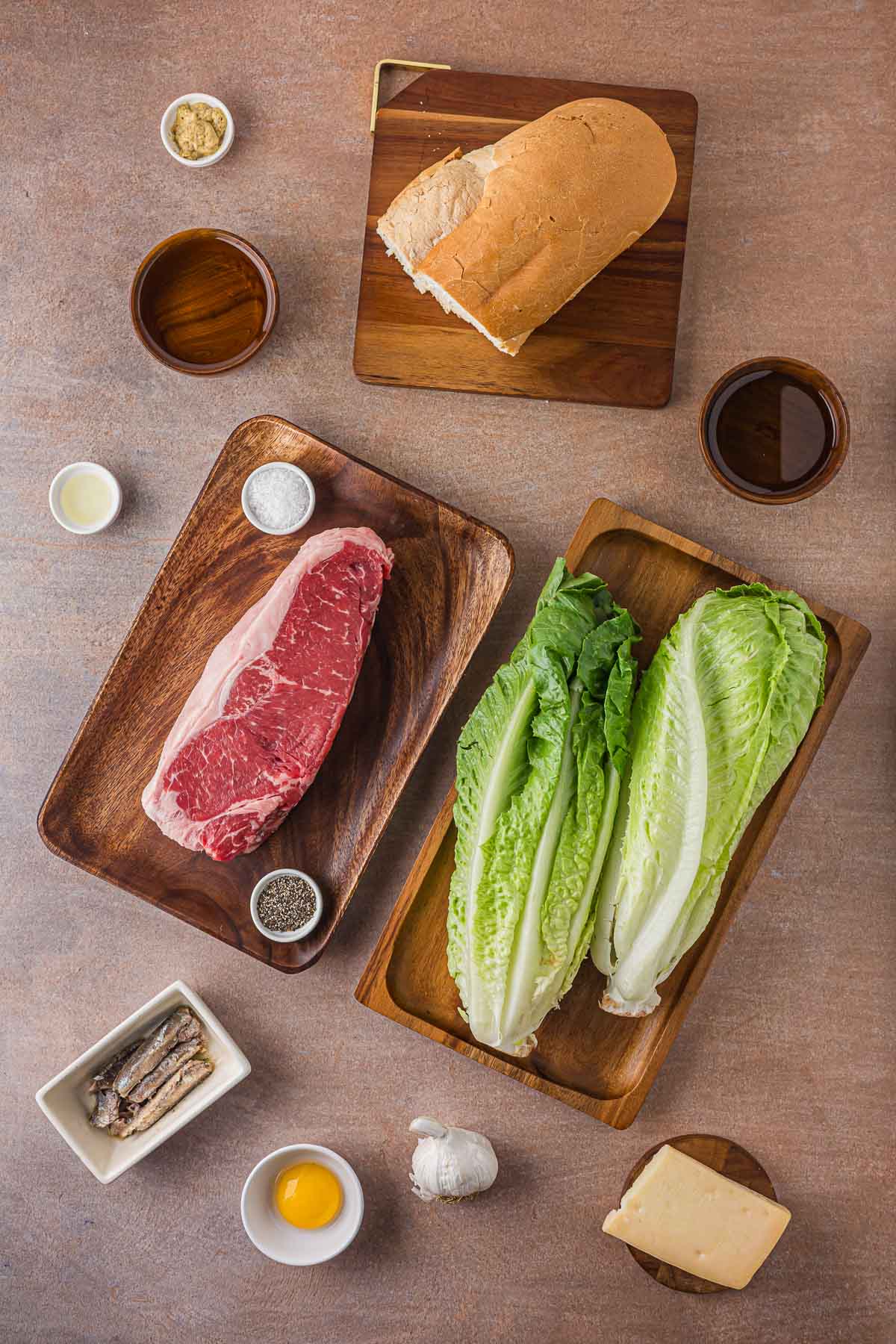 Ingredients for grilled steak Caesar salad: raw New York strip, romaine hearts, Italian bread, anchovies, an egg yolk, garlic, parmesan, lemon, mustard and olive oil.