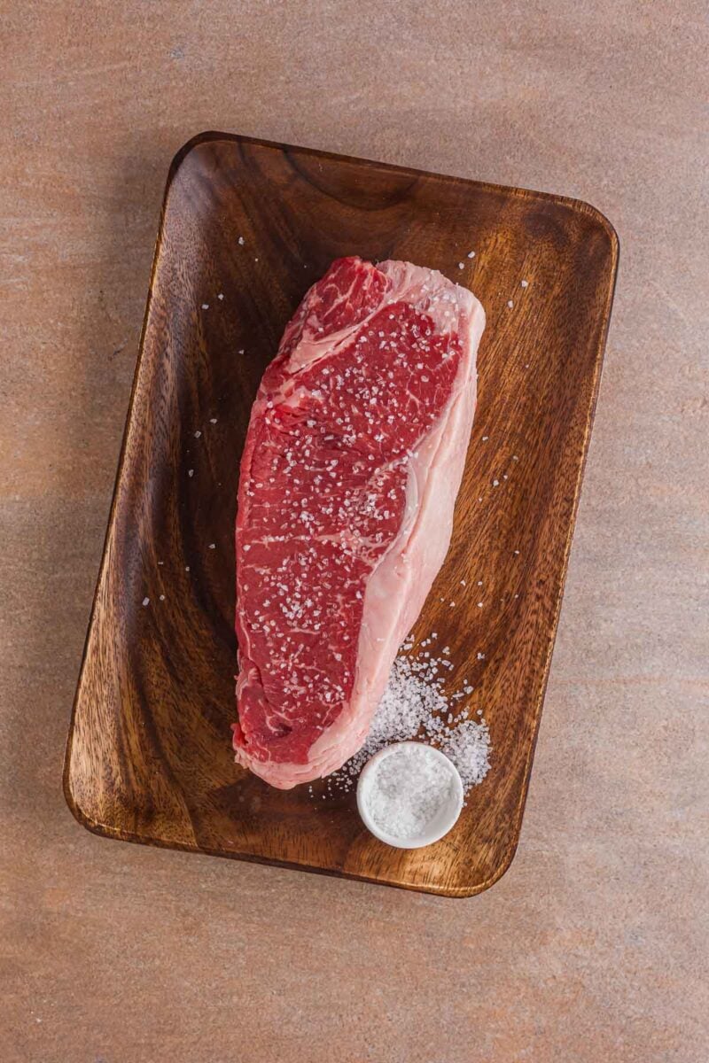 A raw New York strip steak seasoned with coarse salt on a wooden board, with a small bowl of salt beside it.
