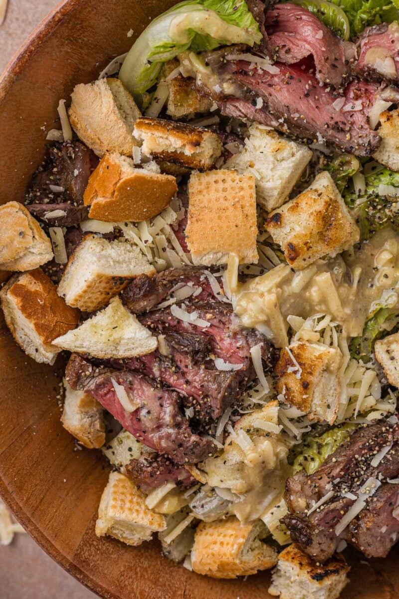 Grilled steak Caesar salad up close, the anchovy garlic dressing pooling between torn croutons, parmesan and pink strip steak.
