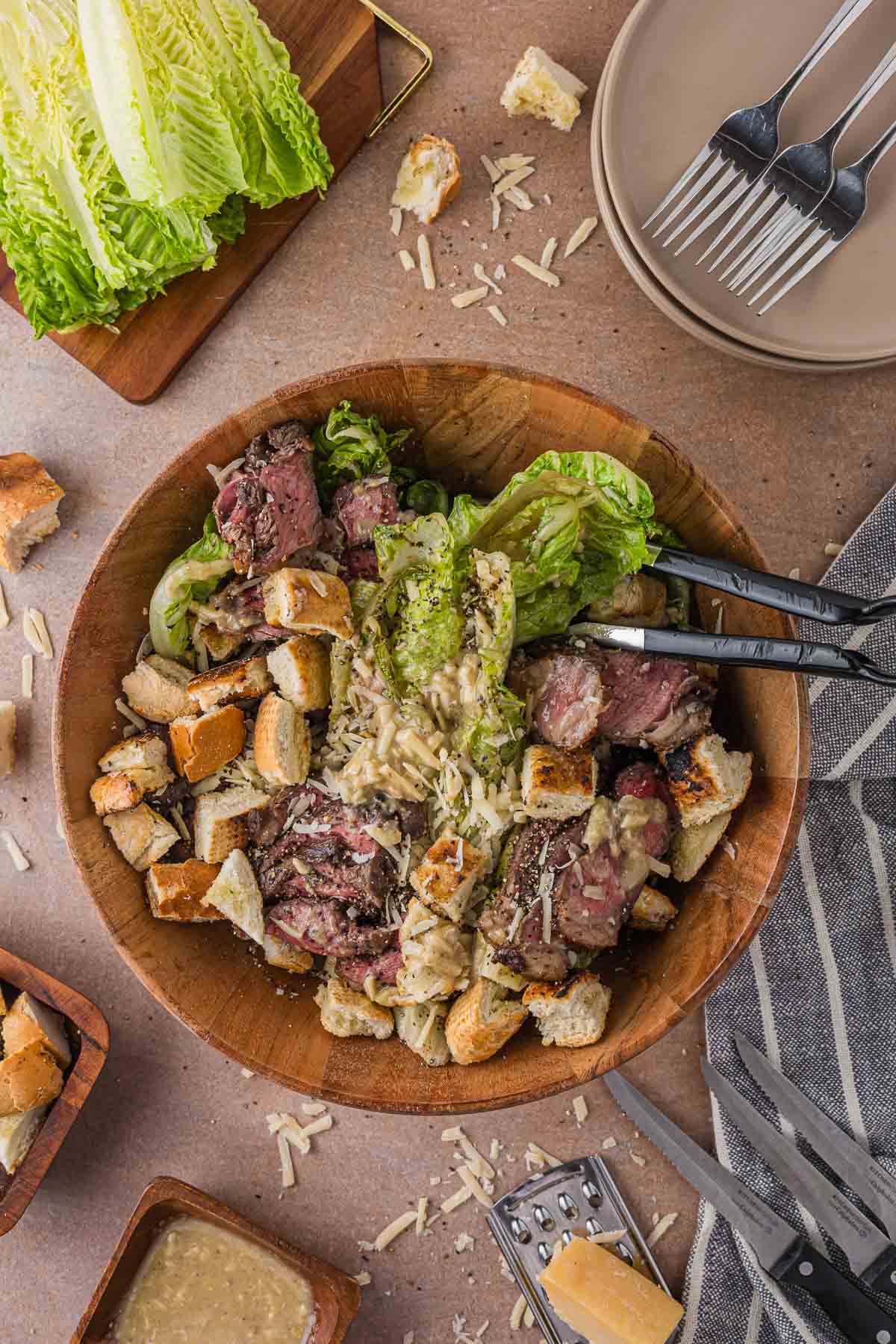 Tongs resting in the wooden bowl of grilled steak Caesar salad, with raw romaine, plates, croutons, dressing and a parmesan wedge staged around it.