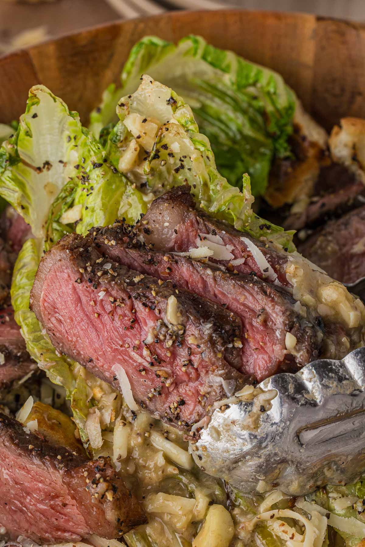 Metal tongs closing around a thick slice of medium-rare grilled New York strip in the Caesar salad bowl.