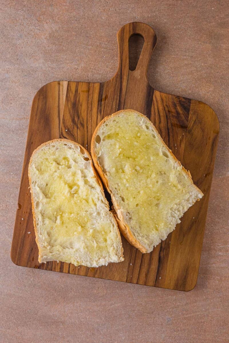 Two thick slices of Italian bread brushed with olive oil on a wooden cutting board, ready to be grilled into Caesar croutons.