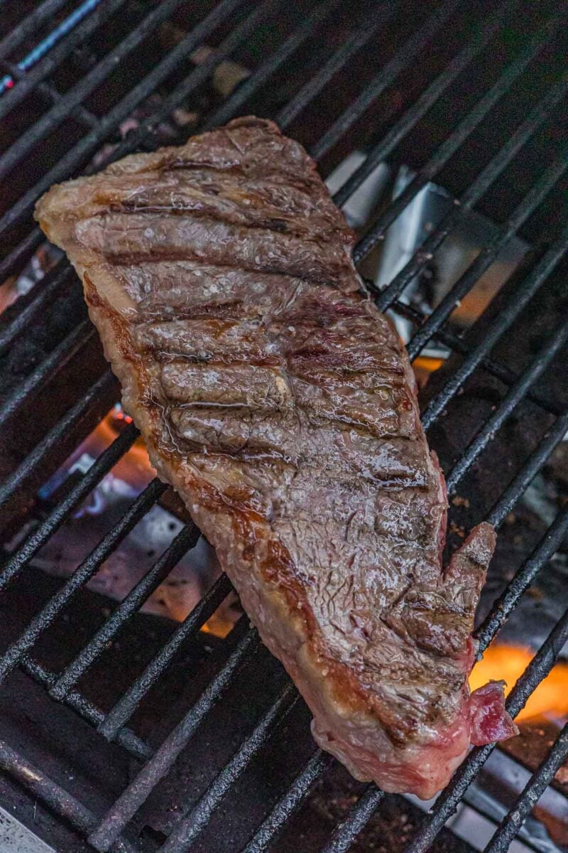 The New York strip flipped on the grill, seared side up with dark crosshatched grill marks across the crust.