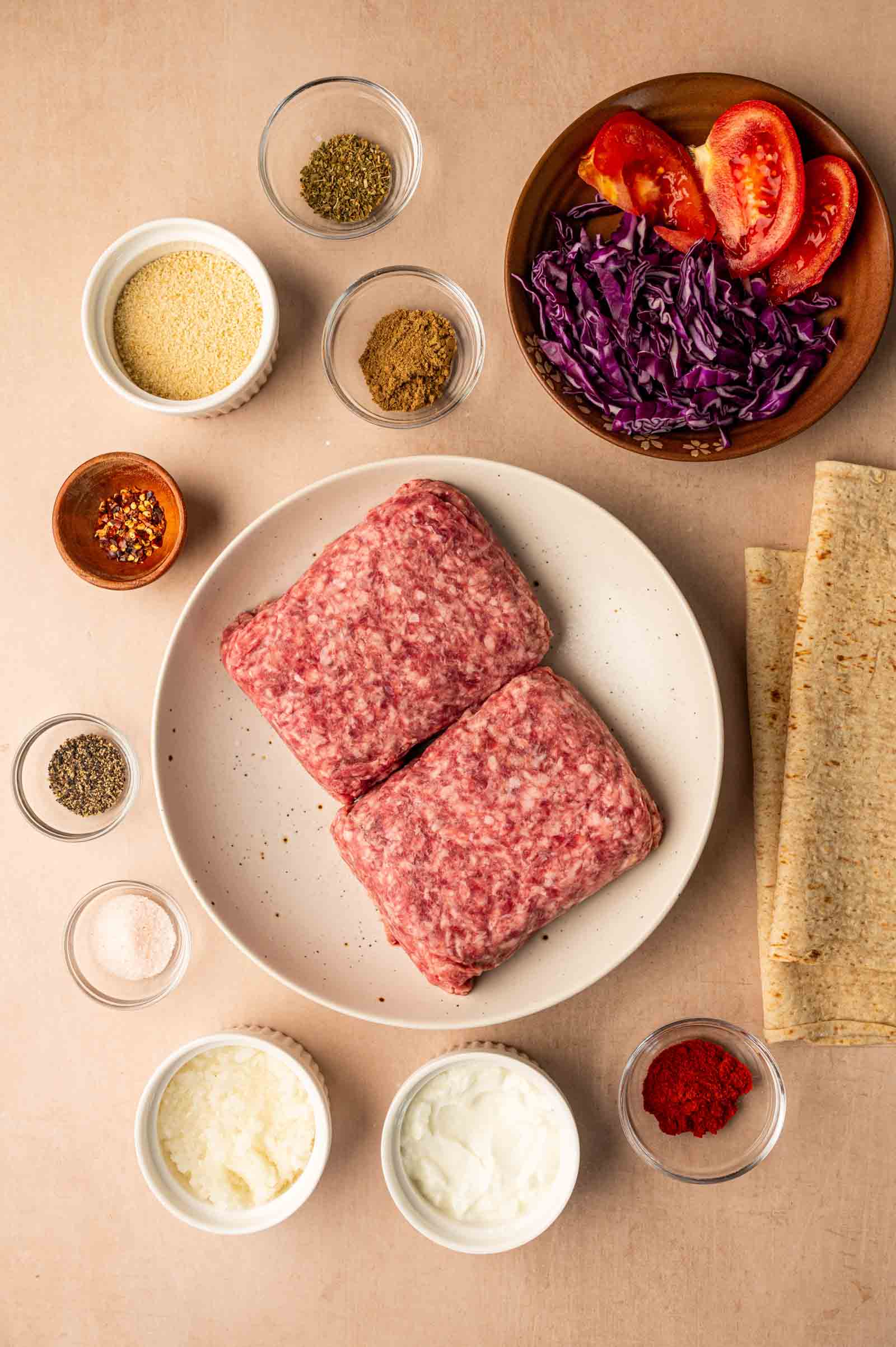 Raw ground lamb on a plate surrounded by spices, tomato wedges, shredded cabbage, flatbreads, yogurt, and sour cream, all arranged on a light surface.