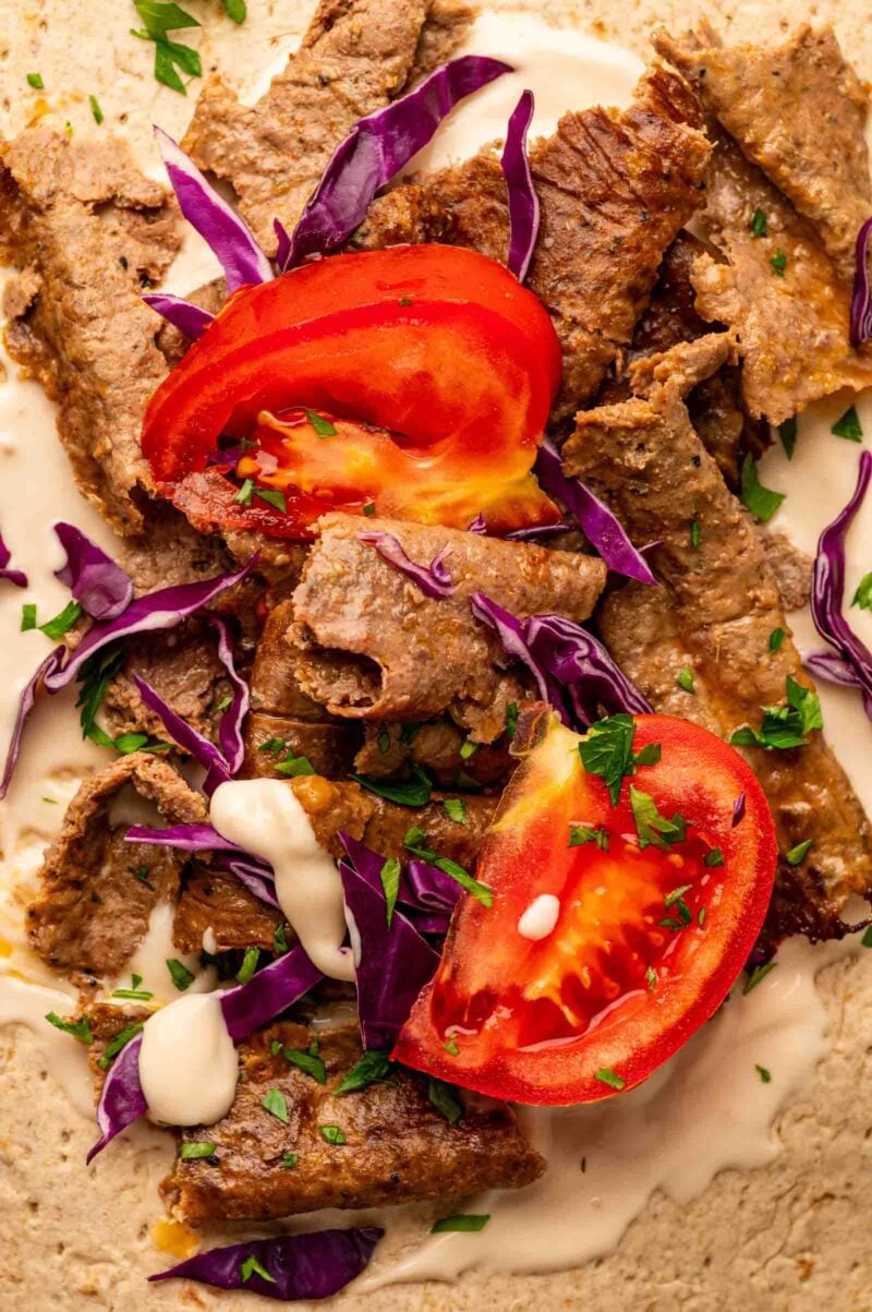 Sliced lamb doner with purple cabbage, tomato wedges, chopped herbs, and white sauce on a flatbread.