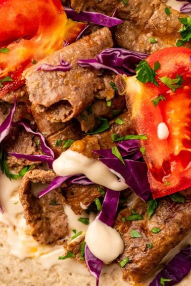 Close-up of sliced lamb, tomato, shredded purple cabbage, chopped herbs, and white sauce on flatbread.