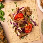 Flatbread topped with sliced lamb, shredded purple cabbage, tomato, chopped parsley, and a drizzle of white sauce, with lemon wedges and herbs on the side.