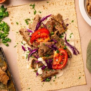 Flatbread topped with sliced lamb, shredded purple cabbage, tomato, chopped parsley, and a drizzle of white sauce, with lemon wedges and herbs on the side.