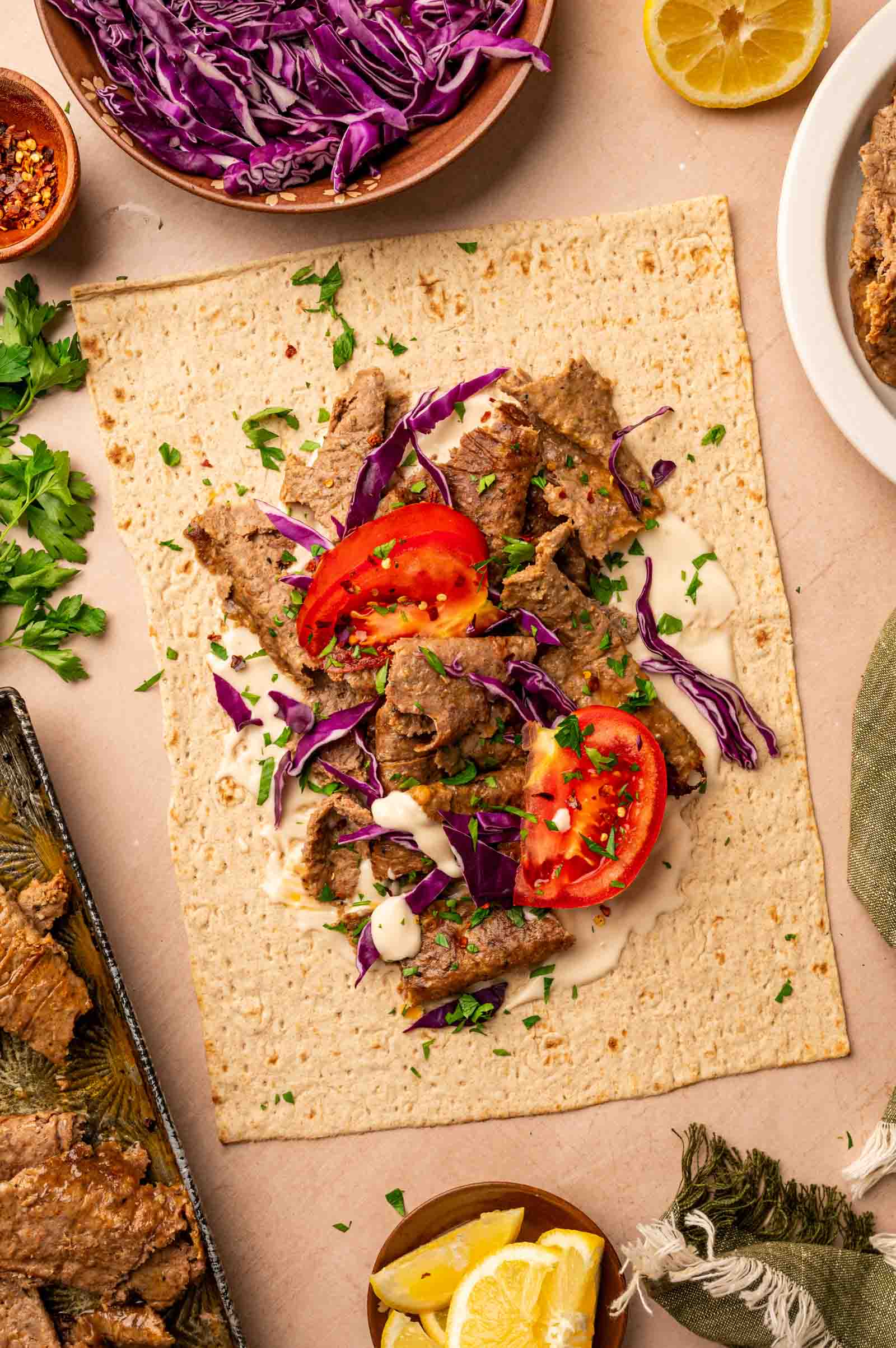 Flatbread topped with sliced lamb, shredded purple cabbage, tomato, chopped parsley, and a drizzle of white sauce, with lemon wedges and herbs on the side.