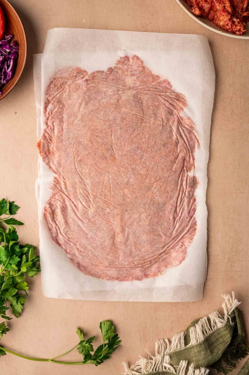 A large piece of raw ground lamb is flattened between two sheets of parchment paper on a beige surface, surrounded by herbs and bowls of ingredients.