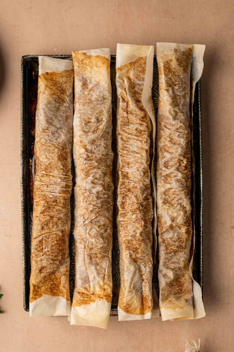 Four rolled, cooker doner lamb meat in a single layer on a rectangular baking tray, viewed from above.