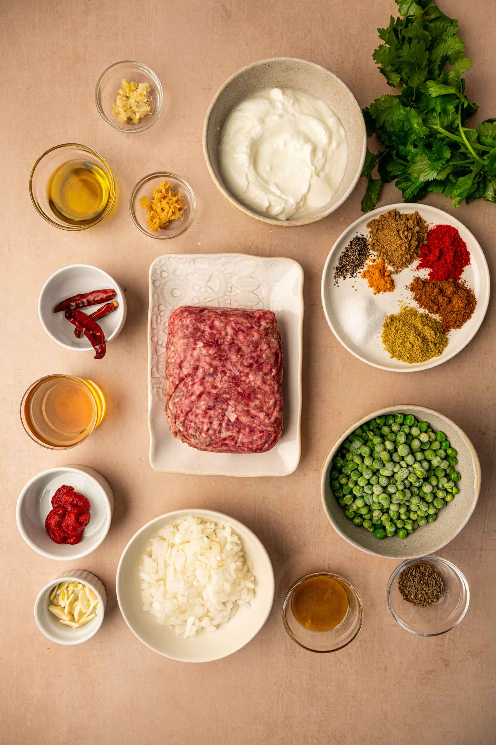 Various keema ingredients including ground lamb, spices, peas, onions, herbs, oil, yogurt, garlic, ginger, lemon zest, and tomato paste arranged in bowls on a beige surface.