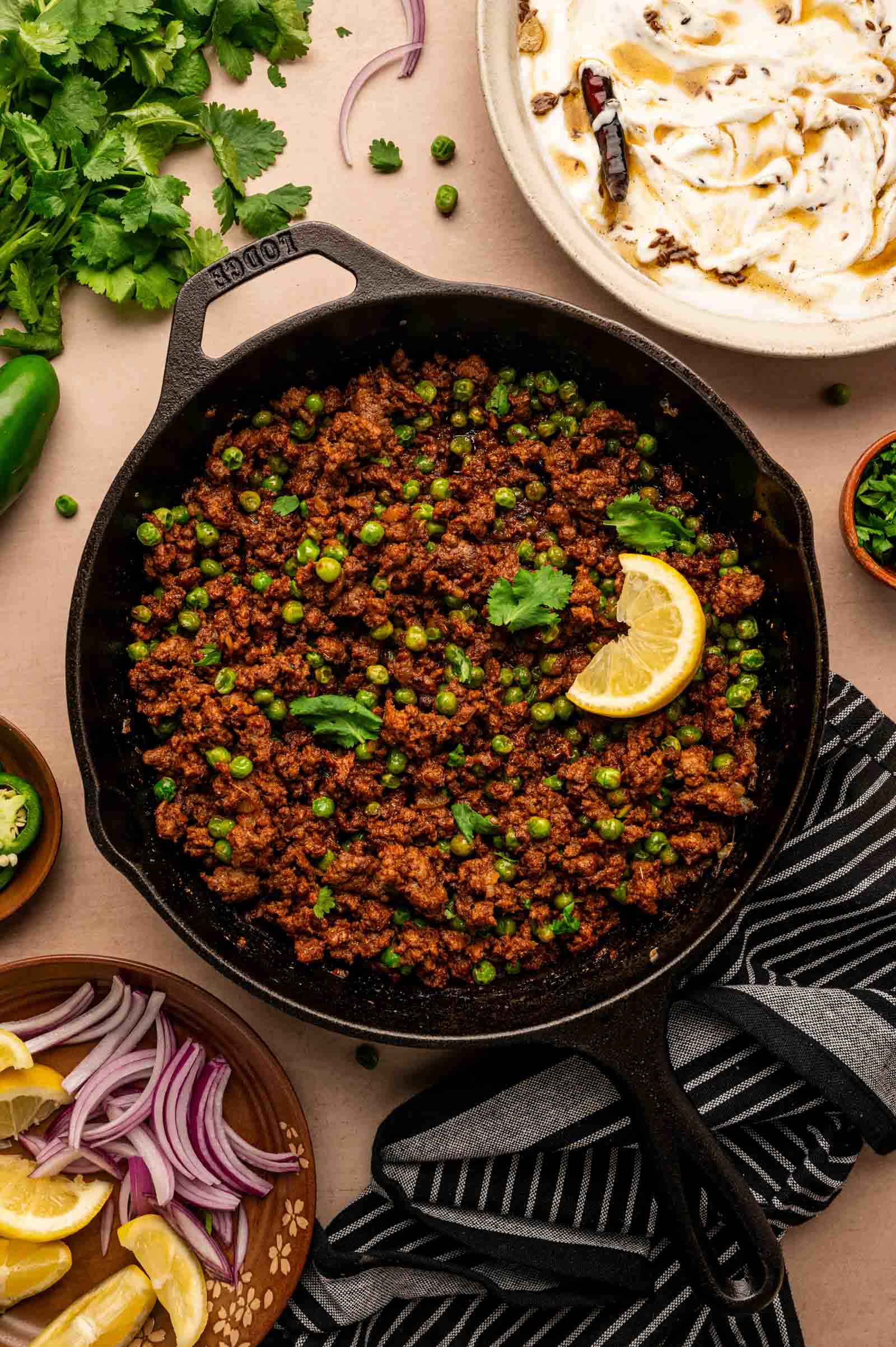 Cast iron skillet filled with ground lamb keema and green peas, garnished with cilantro and a lemon slice, surrounded by lemon wedges, red onion, yogurt, and herbs.